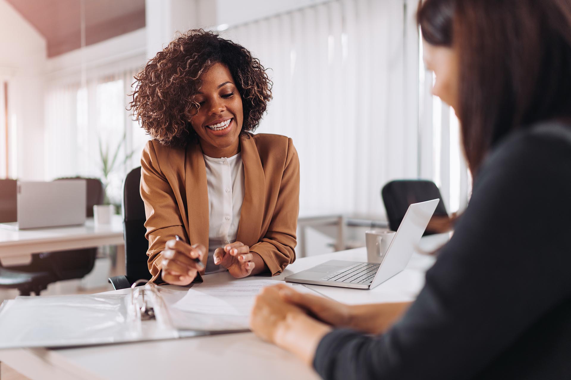  A professional woman in a brown blazer is sitting at a desk, smiling and consulting with a client while pointing at a document.